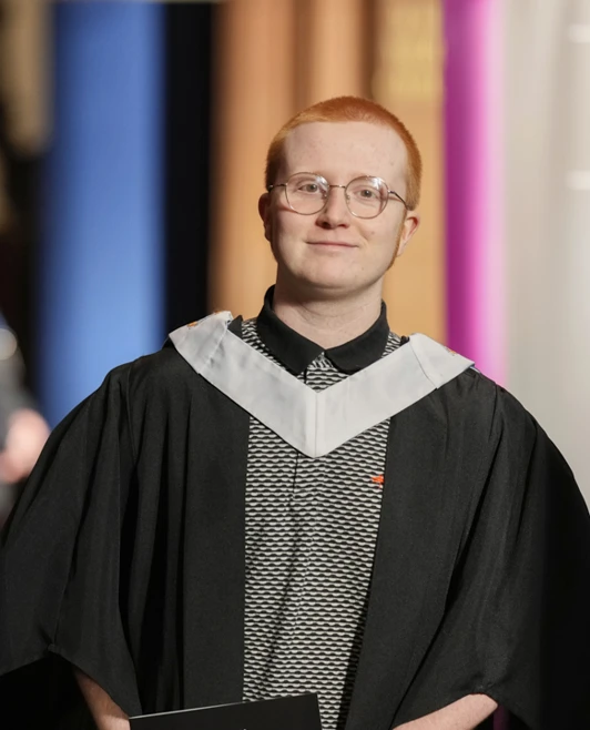 Graduate with glasses smiling in gown and hood during the ceremony Graduate with glasses smiling in gown and hood during the ceremony